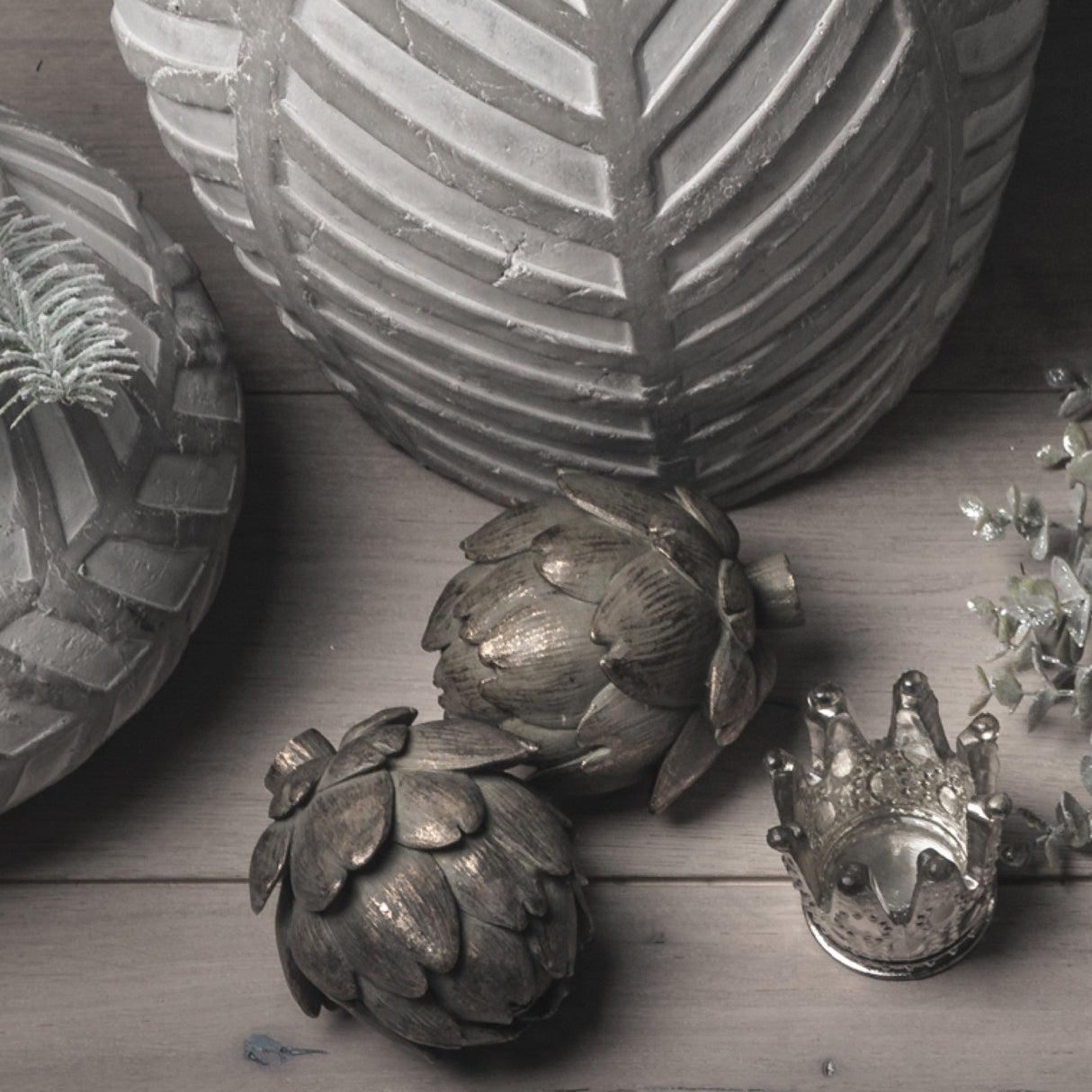Two Hill Interiors antique bronze resin artichoke ornaments grouped with leaf-patterned vases, a small silver crown and eucalyptus on a wooden surface.