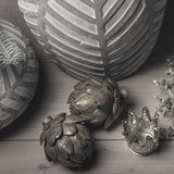 Two Hill Interiors antique bronze resin artichoke ornaments grouped with leaf-patterned vases, a small silver crown and eucalyptus on a wooden surface.
