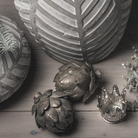 Two Hill Interiors antique bronze resin artichoke ornaments grouped with leaf-patterned vases, a small silver crown and eucalyptus on a wooden surface.