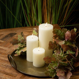 Hill Interiors LED cream wax pillar candles in three staggered heights on a brass tray, styled with autumn oak leaves on a wooden table.