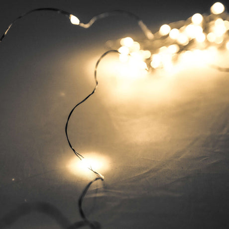 Hill Interiors 100 LED battery micro string lights on a fine silver wire, glowing warm white against a pale draped fabric background.