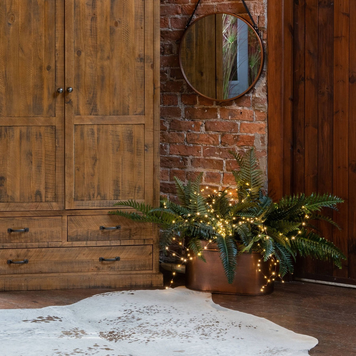 Hill Interiors 100 LED battery micro string lights on silver wire glowing warm white across ferns in a copper planter beside a rustic wood wardrobe.