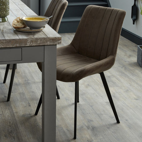 Hill Interiors Draxinby brown faux-leather dining chair with contoured stitched back and slim black tapered legs, set beside a rustic wooden dining table.