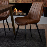 Hill Interiors Draxinby brown faux-leather dining chair with ribbed stitching and black metal legs, set on a dark rug beside a wooden table and lit fireplace.