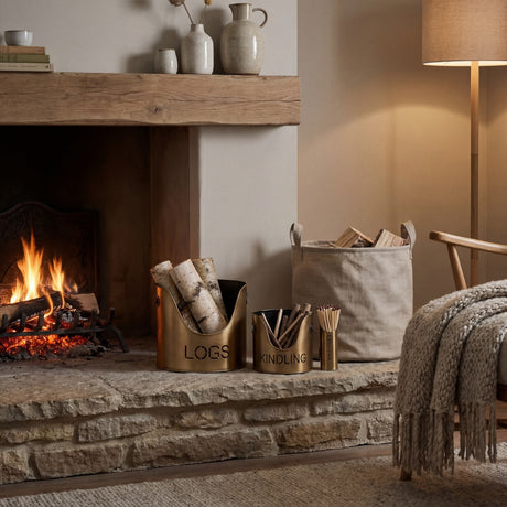 Hill Interiors bronze finish metal log bucket, kindling bucket and matchstick holder set arranged on a stone hearth beside a lit fireplace.