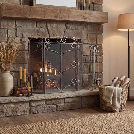 Hill Interiors large fire screen in antique silver metal with scrolled frame and mesh panels, set before a lit stone fireplace with rustic wood mantel.