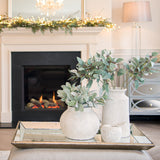 Hill Interiors distressed large mirrored tray with silver wooden trim, styled on an ottoman with stone vases of eucalyptus beside a lit fireplace.