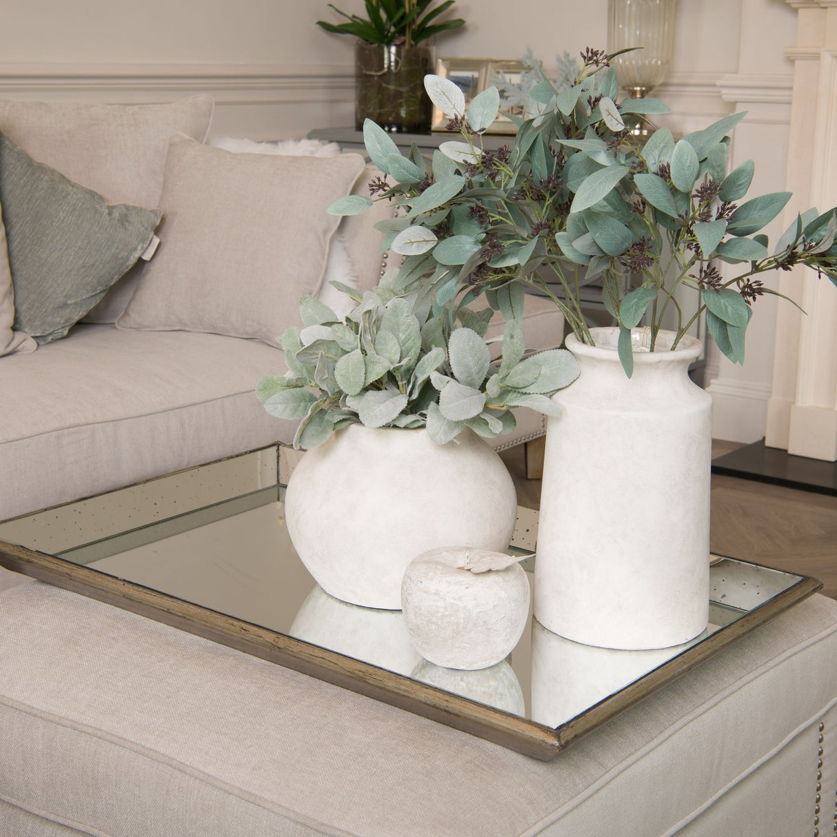 Hill Interiors distressed large mirrored tray with wooden trim on an ottoman, holding two white ceramic vases with eucalyptus foliage in a living room.