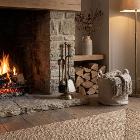 Hill Interiors Ball Topped Companion Set in antique brass beside a stone fireplace with logs stacked, woven basket, and floor lamp in a cosy living room.