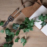 Hill Interiors Faux Ivy Garland 200cm trailing across a wooden surface beside wooden-handled secateurs and an open leather-bound notebook.