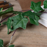 Hill Interiors Faux Ivy Garland 200cm draped across a wooden desk beside an open notebook and wooden-handled tools, showing veined green leaves on purple stems.