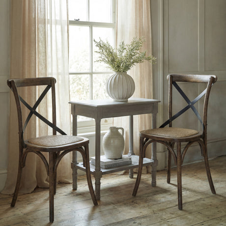 Hill Interiors Cross Back Dining Chairs in brown solid wood with woven seats and metal cross detail, flanking a small table by a curtained window.