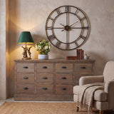 Hill Interiors antique gold hares table lamp with green velvet shade, lit on a wooden drawer sideboard beside a large Roman numeral wall clock.