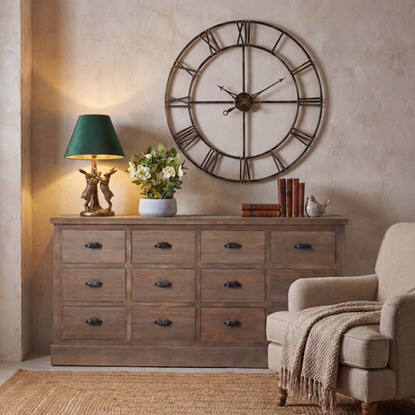Hill Interiors antique gold hares table lamp with green velvet shade, lit on a wooden drawer sideboard beside a large Roman numeral wall clock.