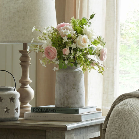 Hill Interiors urn-shaped stone-effect ceramic vase in beige holding pink roses and white blooms, styled on books beside a lamp by a sunlit window.