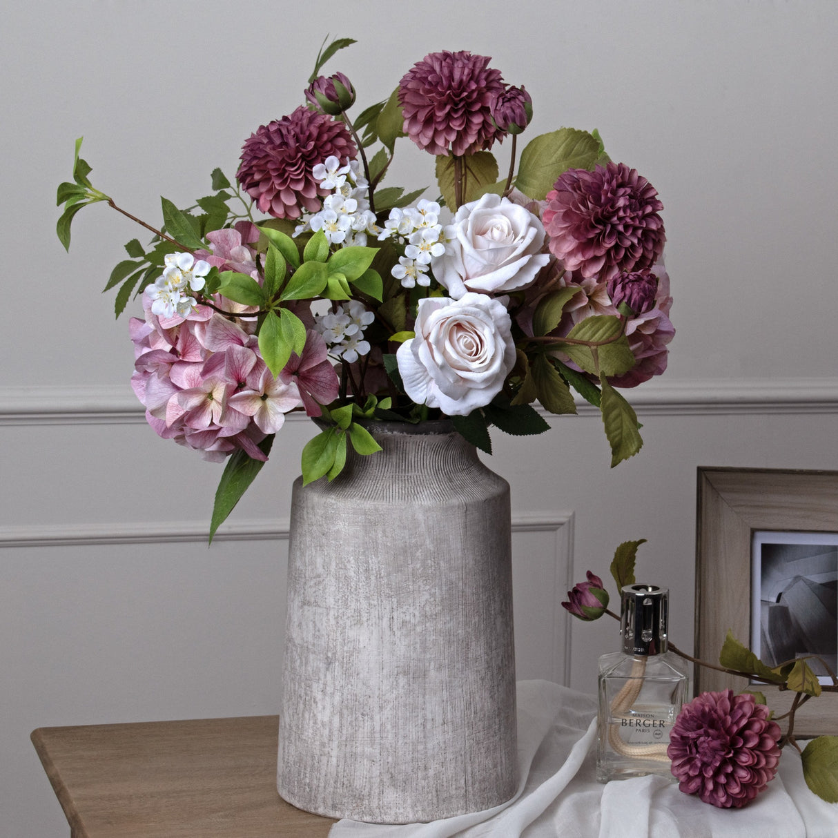 Hill Interiors urn-shaped stone-effect ceramic vase in beige, holding pink roses, mauve dahlias and hydrangeas on a wooden console against a pale wall.
