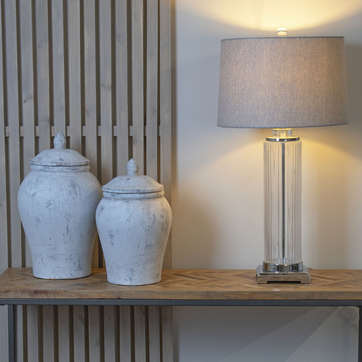 Two Hill Interiors stone ceramic ginger jar vases in beige with domed lids on a wooden console, beside a ribbed glass table lamp with grey shade.