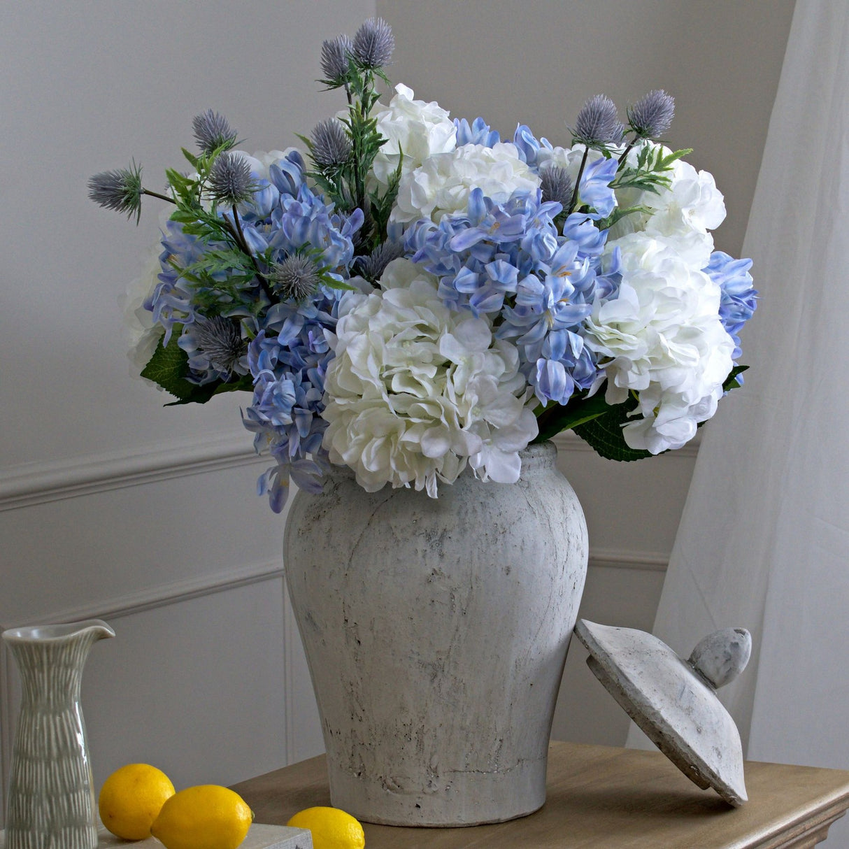 Hill Interiors Stone Ginger Jar ceramic vase in beige, styled with white and blue hydrangeas and thistles, lid resting beside it on a wooden console.