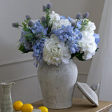 Hill Interiors Stone Ginger Jar ceramic vase in beige, styled with white and blue hydrangeas and thistles, lid resting beside it on a wooden console.