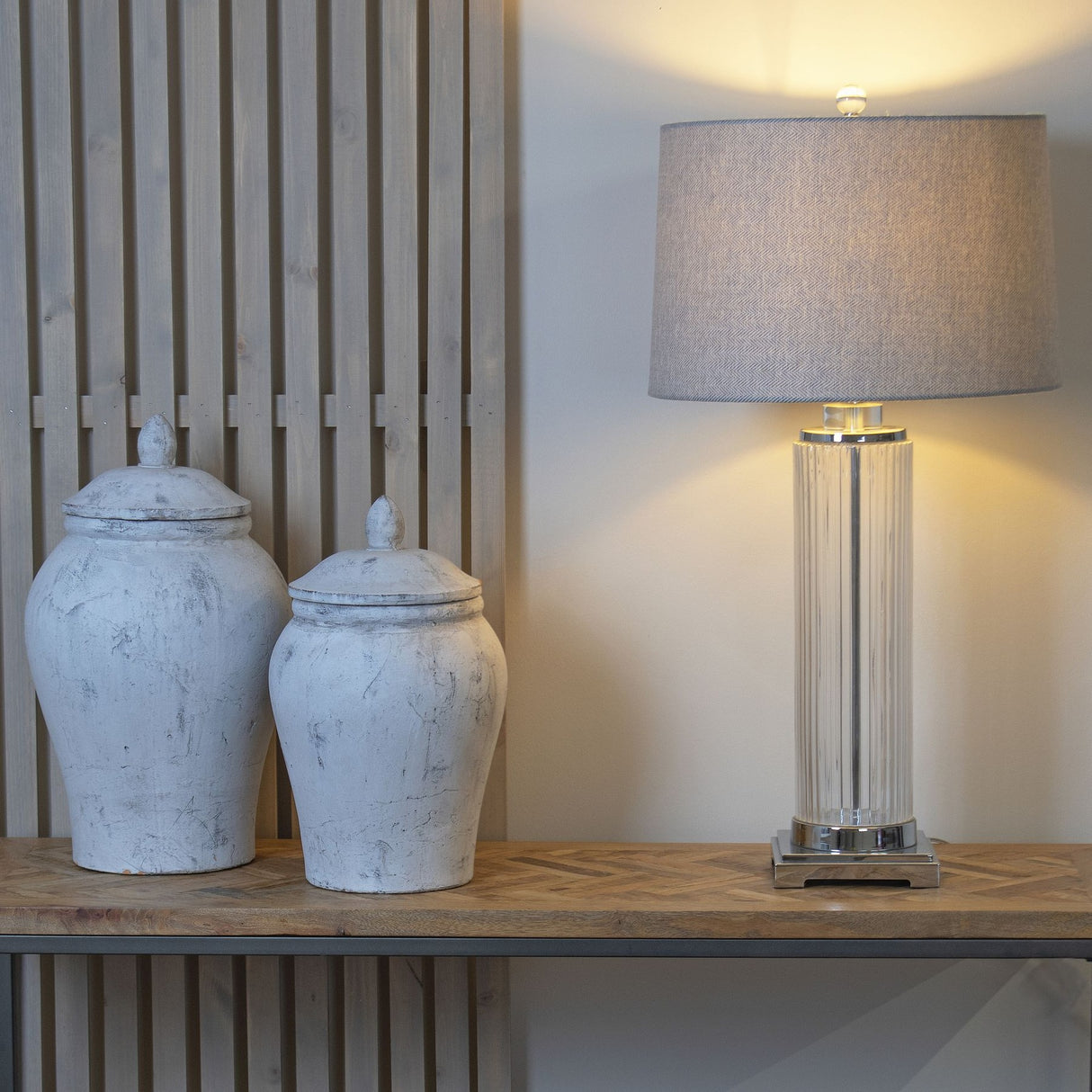 Two Hill Interiors Stone Ginger Jar ceramic vases in beige with domed lids on a wooden console beside a fluted glass table lamp with grey shade.