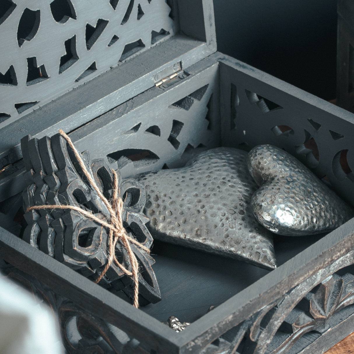 Hill Interiors silver ceramic hearts with dimpled glaze resting inside an open carved dark grey wooden box alongside a tied ornate cut-out panel.