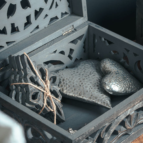 Hill Interiors silver ceramic hearts with dimpled glaze resting inside an open carved dark grey wooden box alongside a tied ornate cut-out panel.