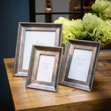 Hill Interiors Convex Matt Silver Photo Frames in three sizes displayed on a wooden table beside green flowers against a dark blue backdrop.