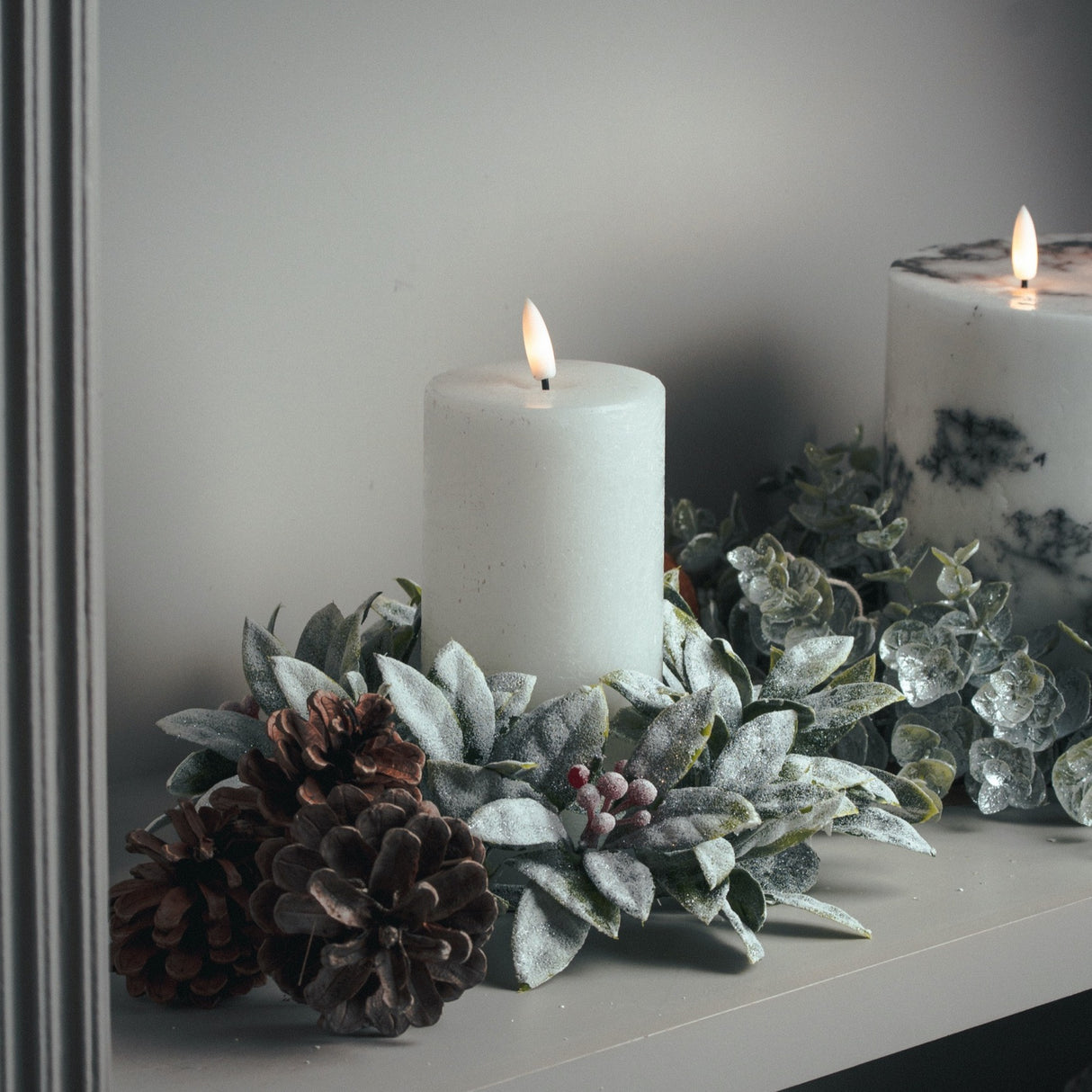 Hill Interiors Natural Glow LED wax pillar candles in beige on a mantel with frosted foliage, red berries and pine cones.
