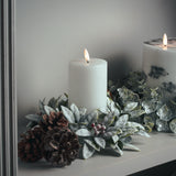 Hill Interiors Natural Glow LED wax pillar candles in beige on a mantel with frosted foliage, red berries and pine cones.