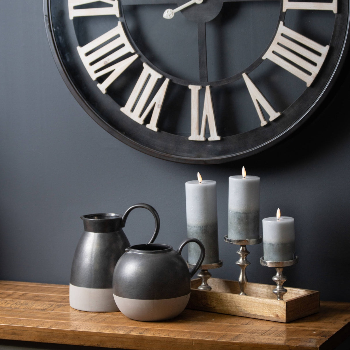 Hill Interiors Natural Glow Stone LED wax pillar candles in beige on silver holders atop a wooden sideboard beside black jugs and a Roman numeral clock.