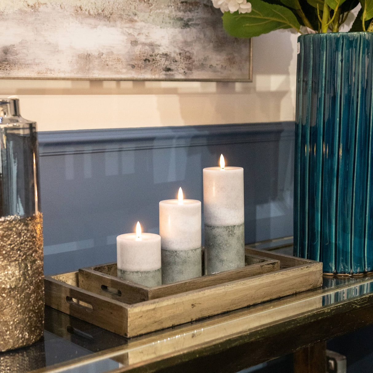 Hill Interiors Natural Glow Stone LED wax pillar candles in beige, grouped in three heights on a wooden tray beside a teal vase on a mirrored console.