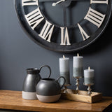 Hill Interiors Natural Glow Stone LED pillar candles in beige on silver holders atop a wooden tray beside matte black jugs, below a Roman numeral clock.