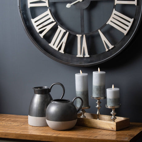 Three lit Hill Interiors Natural Glow stone-effect LED pillar candles on silver holders on a wooden sideboard, beside black jugs under a Roman numeral clock.