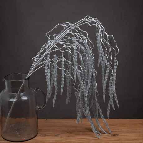 Hill Interiors Festive Faux Amaranthus Stem 130cm in silver, with trailing glittered tassels arranged in a clear glass jug on a wooden surface.