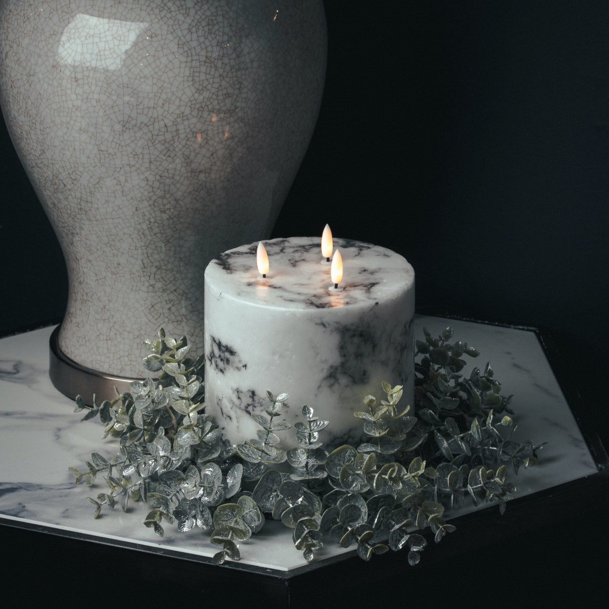 Hill Interiors frosted eucalyptus wreath in white encircling a lit marble-effect pillar candle on a mirrored tray beside a crackle-glaze vase.