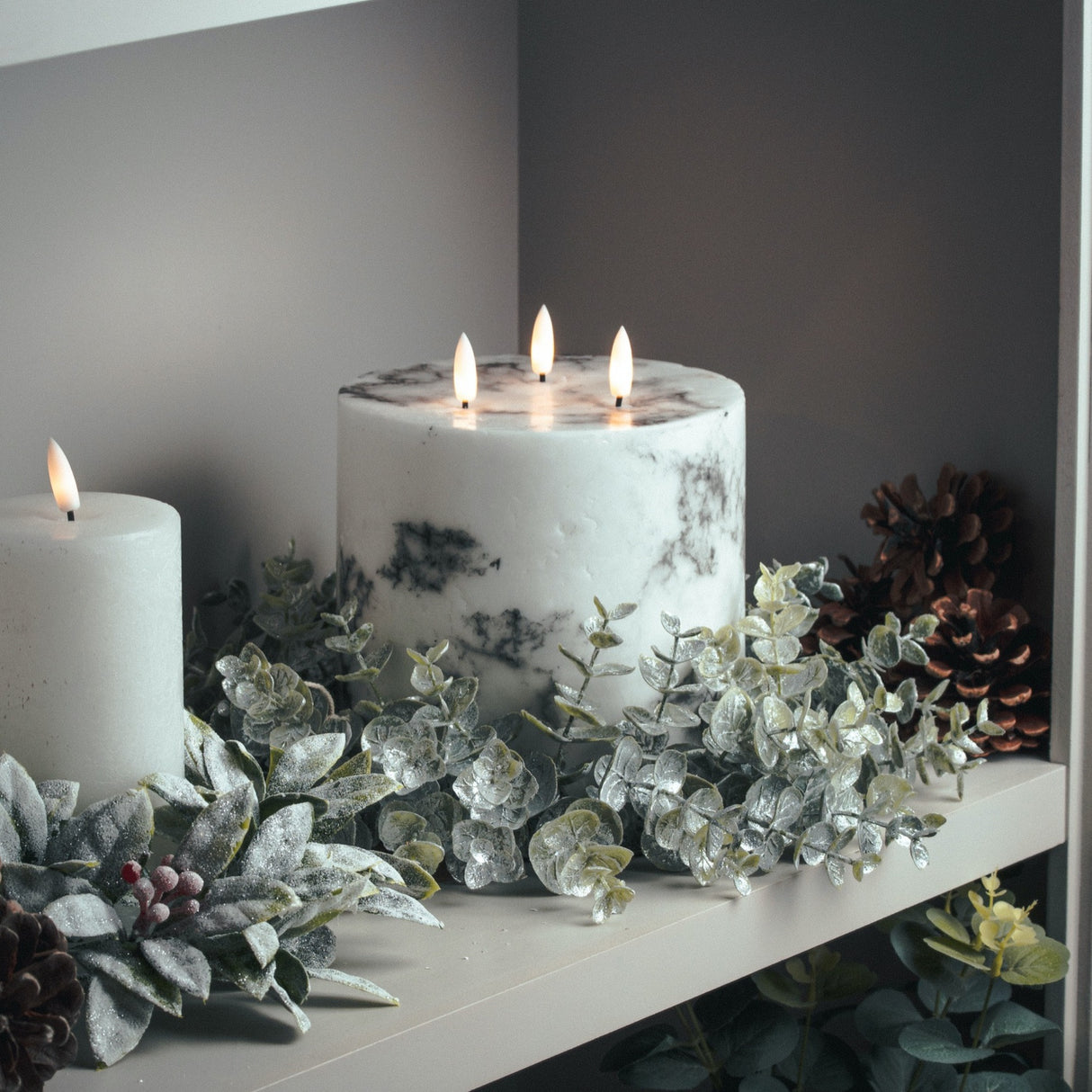 Hill Interiors Large Frosted Eucalyptus Candle Wreath in white encircling a marbled pillar candle beside a white candle and pine cones on a shelf.