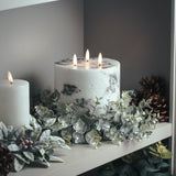 Hill Interiors Large Frosted Eucalyptus Candle Wreath in white encircling a marbled pillar candle beside a white candle and pine cones on a shelf.