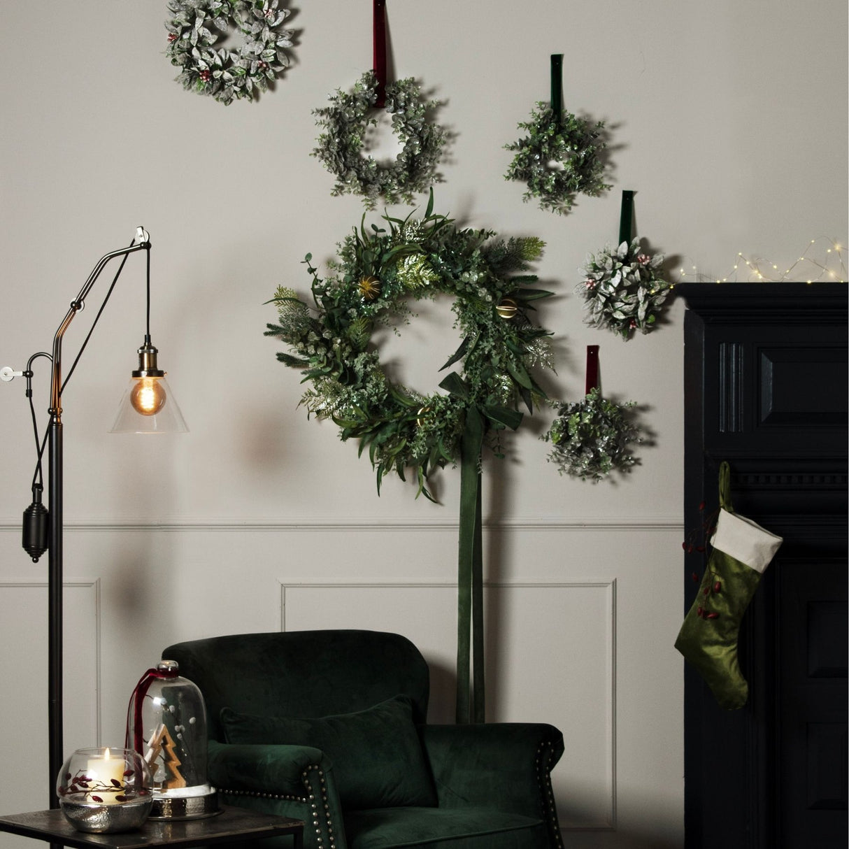 Hill Interiors frosted eucalyptus wreaths in white and green displayed on a pale panelled wall above a green velvet armchair with floor lamp and stocking.