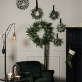 Hill Interiors frosted eucalyptus wreaths in white and green displayed on a pale panelled wall above a green velvet armchair with floor lamp and stocking.