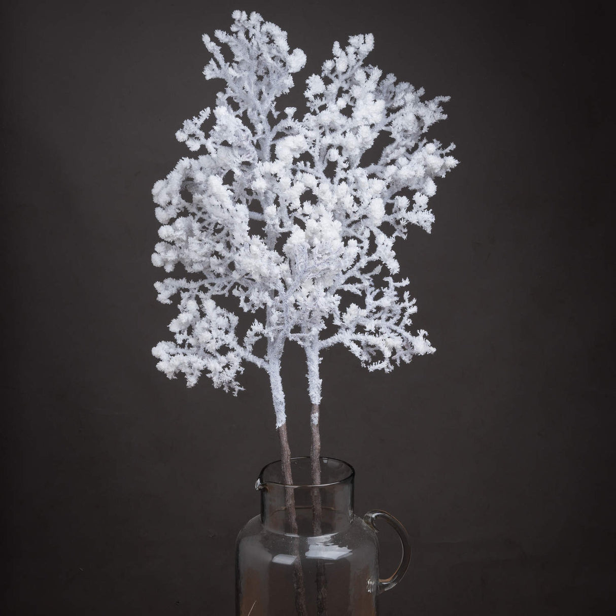 Hill Interiors Snowy Faux Branch 110cm with frosted white moulded plastic twigs displayed upright in a clear glass jug against a dark background.