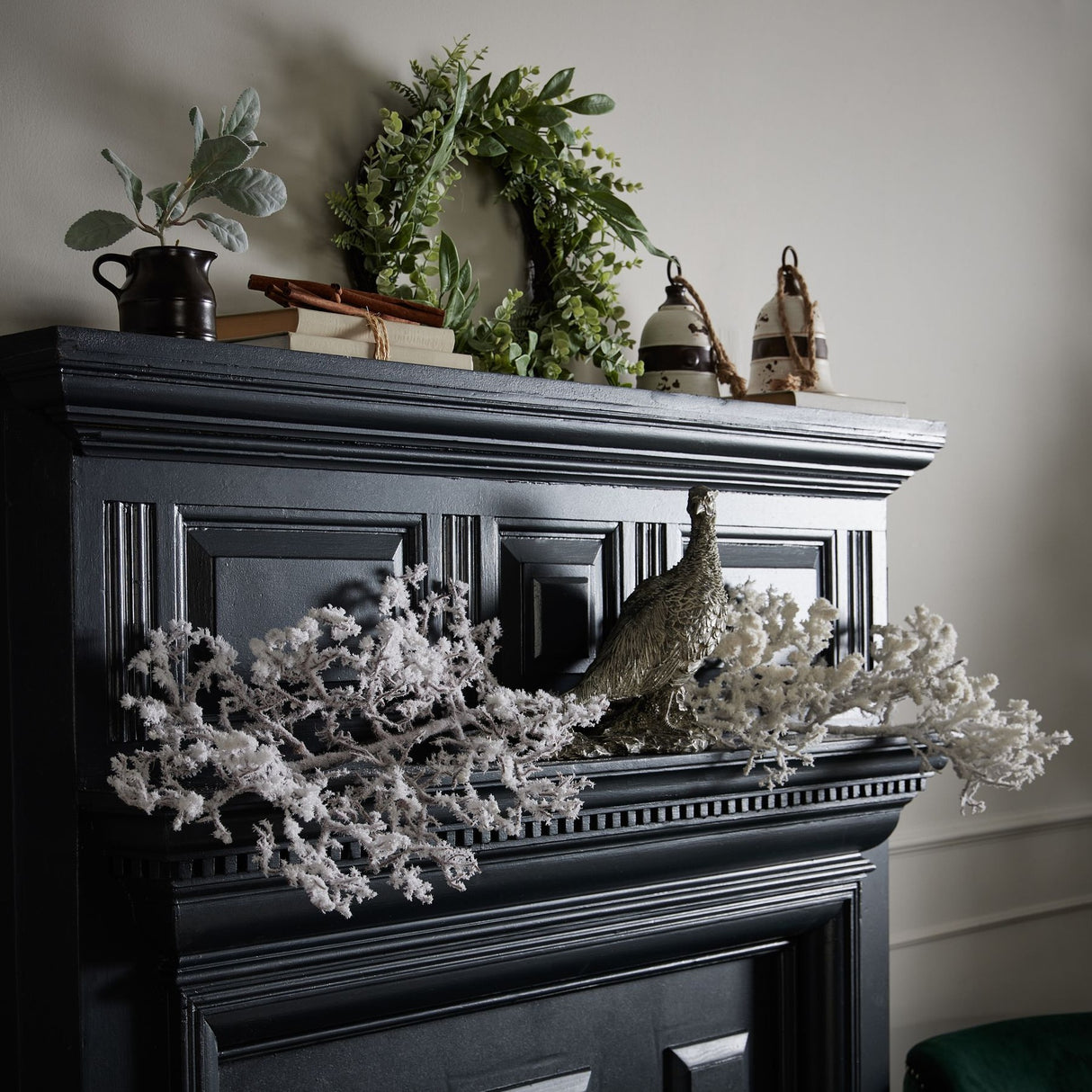 Hill Interiors Snowy Faux Branch 110cm displayed on a black panelled mantelpiece with a pheasant figurine, greenery wreath, ceramic jug and books.