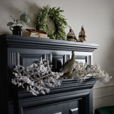 Hill Interiors Snowy Faux Branch 110cm displayed on a black panelled mantelpiece with a pheasant figurine, greenery wreath, ceramic jug and books.
