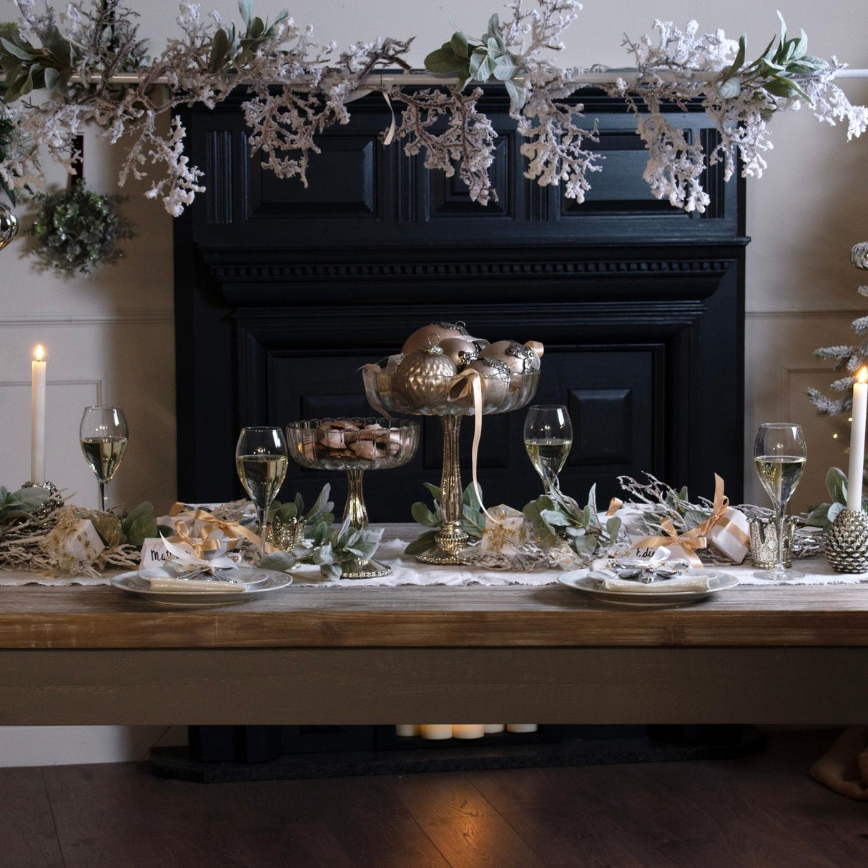 Hill Interiors Snowy Faux Garland draped along a black mantelpiece above a festive dining table set with champagne glasses and candles.