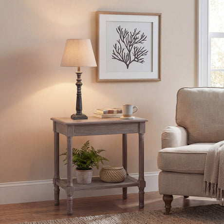 Hill Interiors Ondelle turned wooden column table lamp in warm brown with a tapered shade, lit on a side table beside a cream armchair and framed coral print.