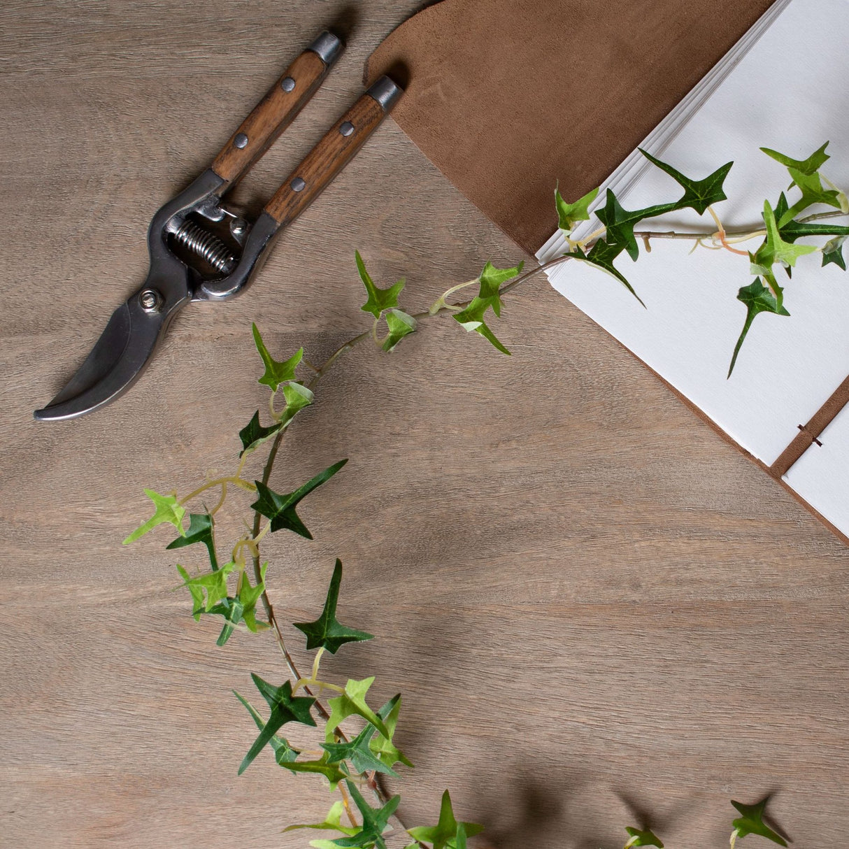 Hill Interiors small faux ivy garland in green trailing across a wooden surface beside wooden-handled pruning shears and an open leather-bound journal.