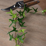 Hill Interiors Small Faux Ivy Garland in green trailing across a wooden surface beside wood-handled pruning shears.