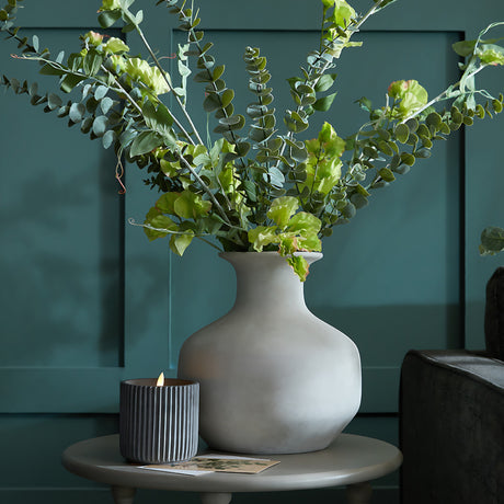 Hill Interiors Squat Stone Ceramic Vase in beige with eucalyptus stems on a round side table beside a ribbed grey candle against a teal panelled wall.
