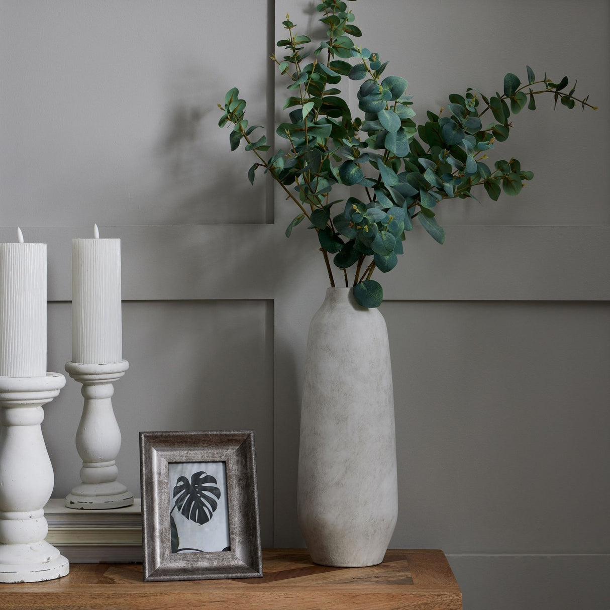 Hill Interiors Large Tall Ceramic Vase in beige holding eucalyptus stems on a wooden console beside white candle holders and a framed print.