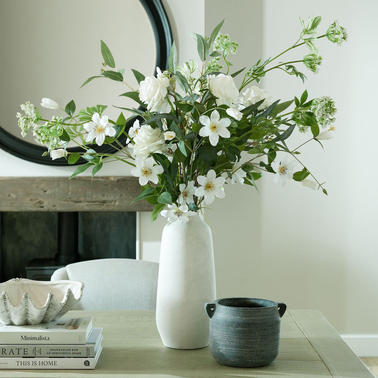 Hill Interiors Large Tall Ceramic Vase in beige holding white flowers and greenery on a wooden console beside a round black mirror and dark handled pot.