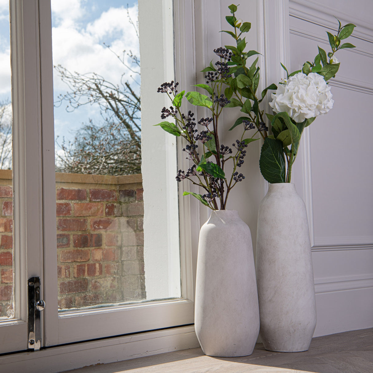 Hill Interiors Tall Ceramic Vase in beige with white hydrangea and berry stems, paired with a smaller vase on a sunlit window sill.